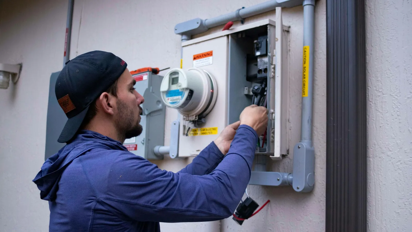 Technician in a cap working on an electrical meter outside a building